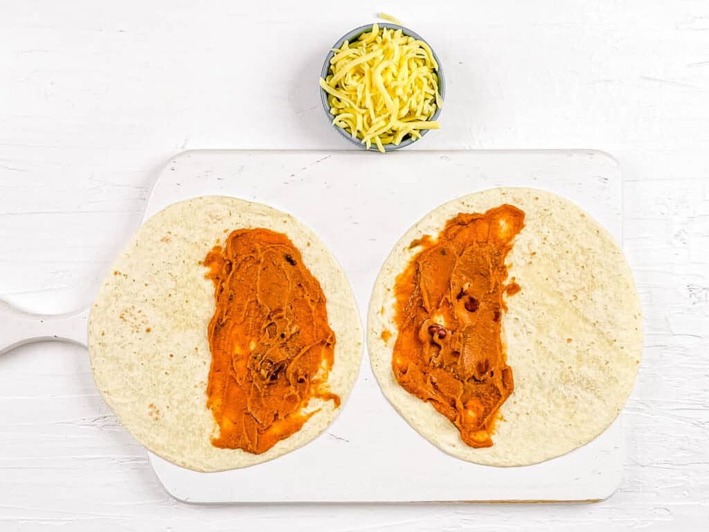 Tortillas topped with refried beans on a cutting board.
