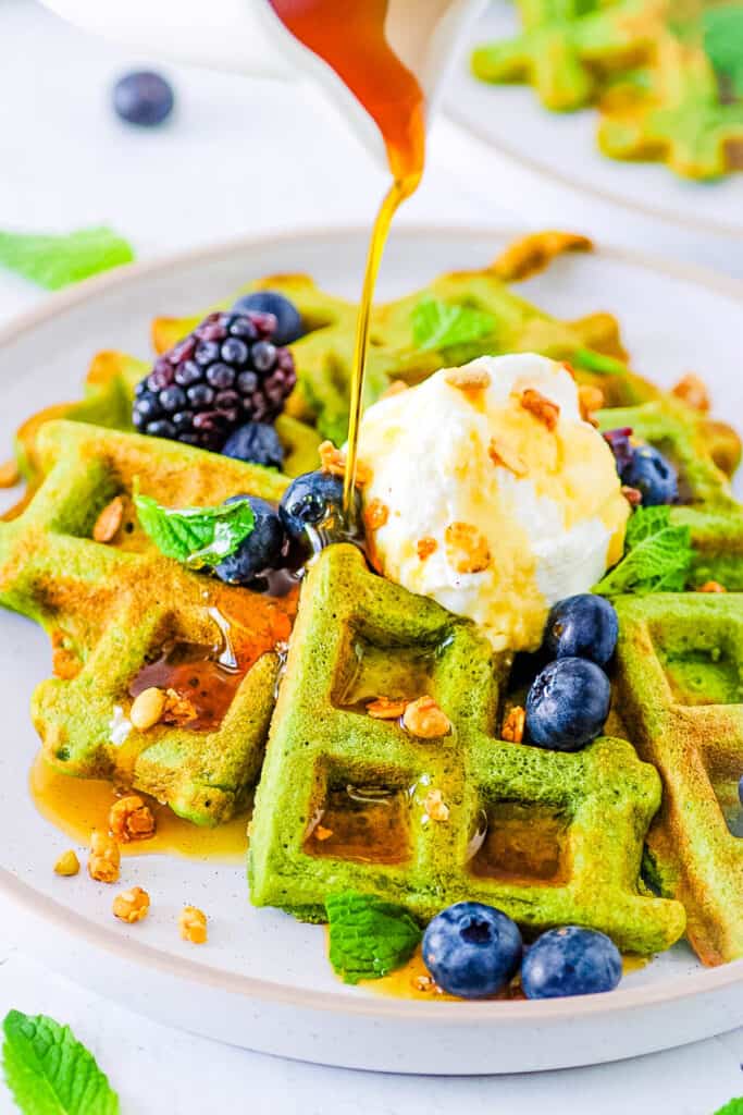 Healthy green waffles served on a white plate with fresh berries, whipped cream and maple syrup.