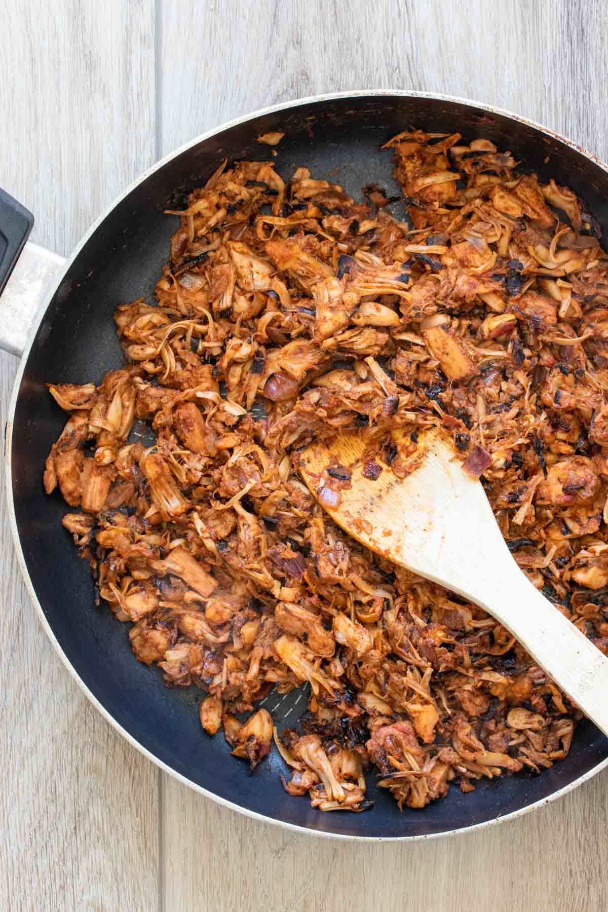 Browning jackfruit with salsa in a pan being mixed by a wooden spoon.