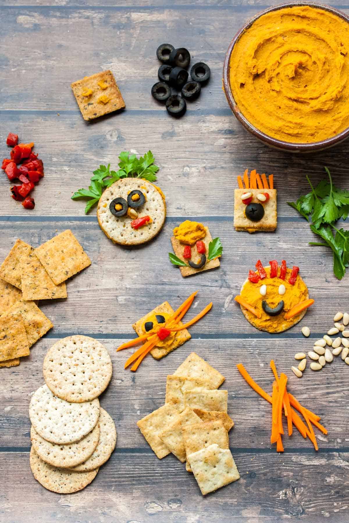 Top view of crackers on a wooden surface with veggies and hummus made into faces