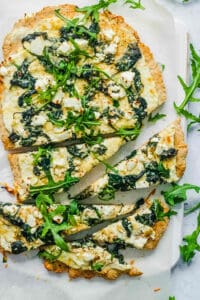 An overhead shot of sliced spinach pizza on a white cutting board.