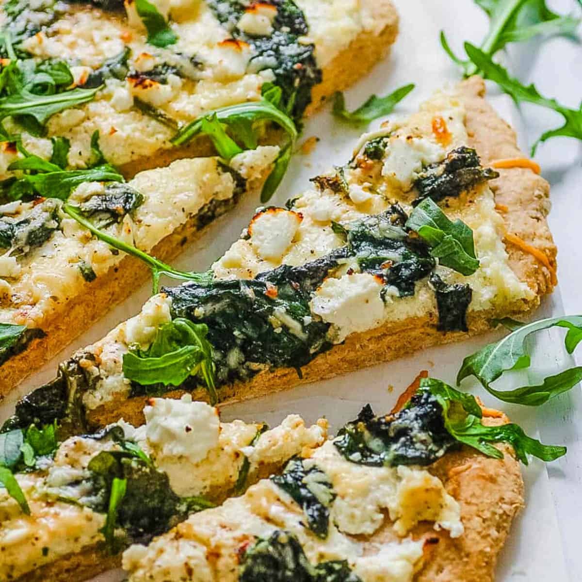 Up close s،t of spinach pizza with ricotta and feta cheese sliced on a white platter.