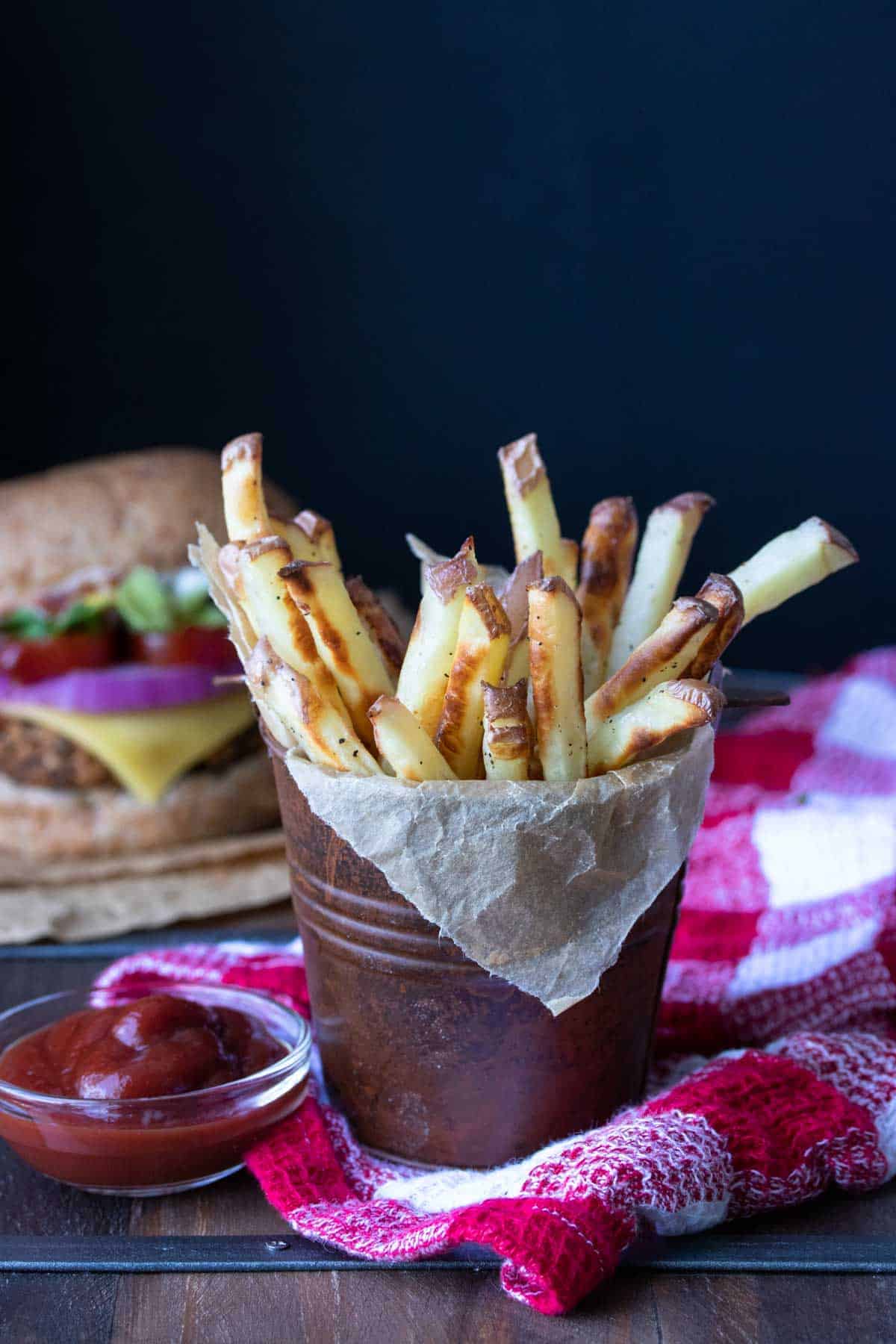 Vegan oven baked french fries in a little tin next to ketchup with a burger in the background.