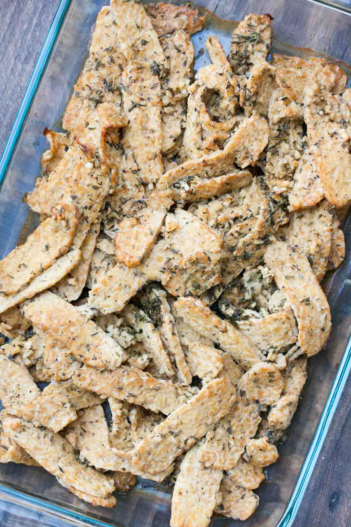 A glass baking dish with slices of tempeh covered in dried herbs and seasoning.