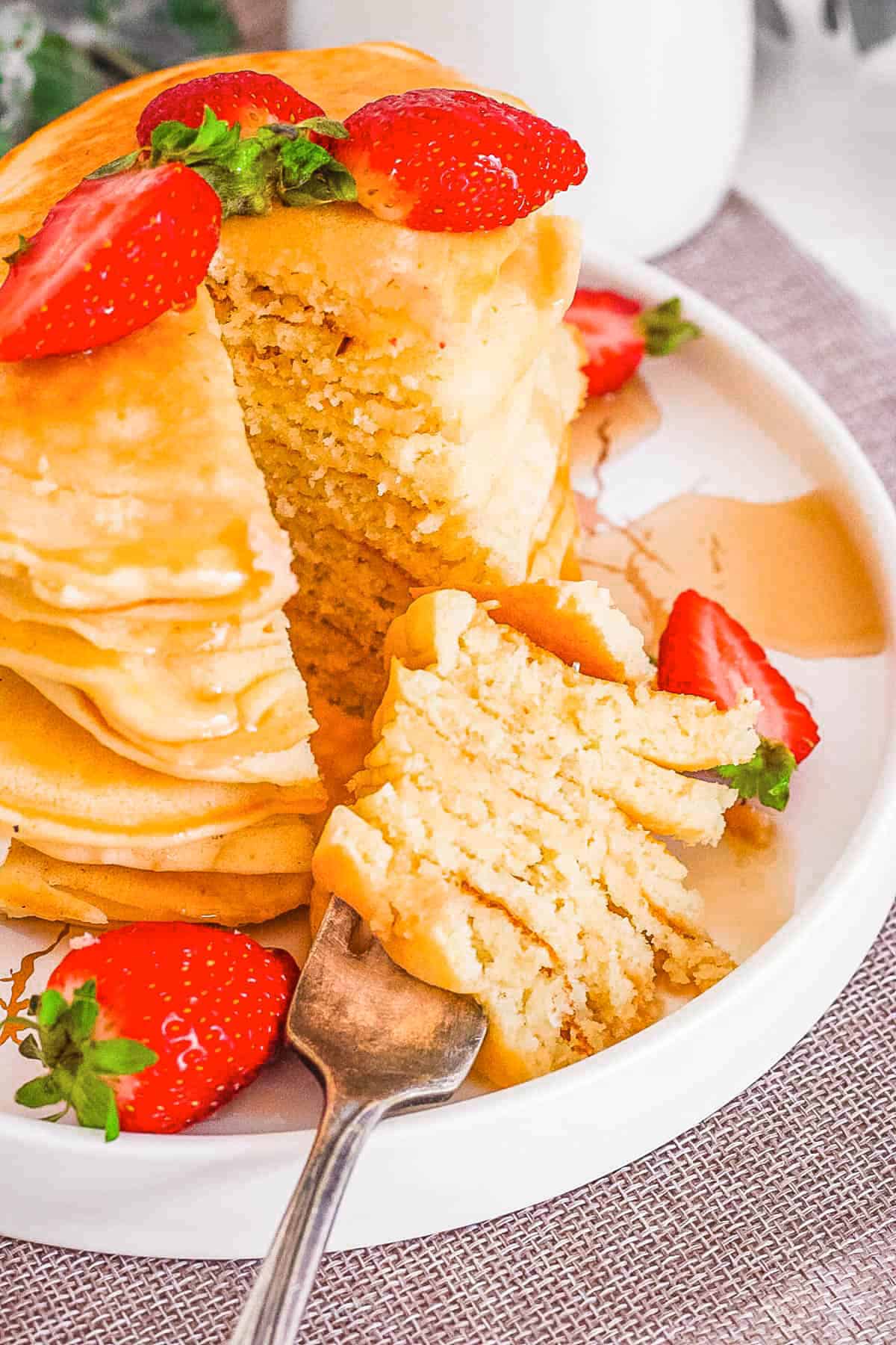 A tall stack of almond milk pancakes drizzled with maple syrup on a white plate, served with strawberries on the side.