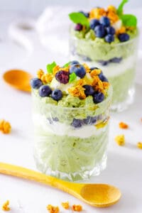 Matcha overnight oats recipe served in a tall glass topped with berries, granola, maple syrup, and mint leaves.