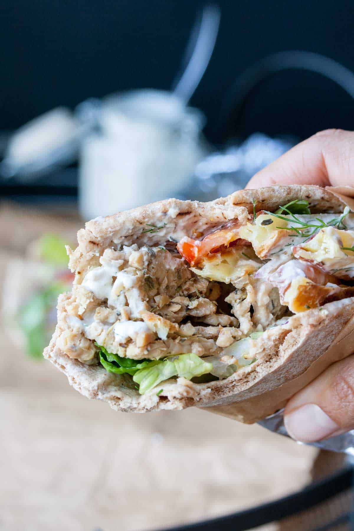 Hand holding a vegan tempeh gyro rolled up in pita bread with veggies, with a bite taken out of it.