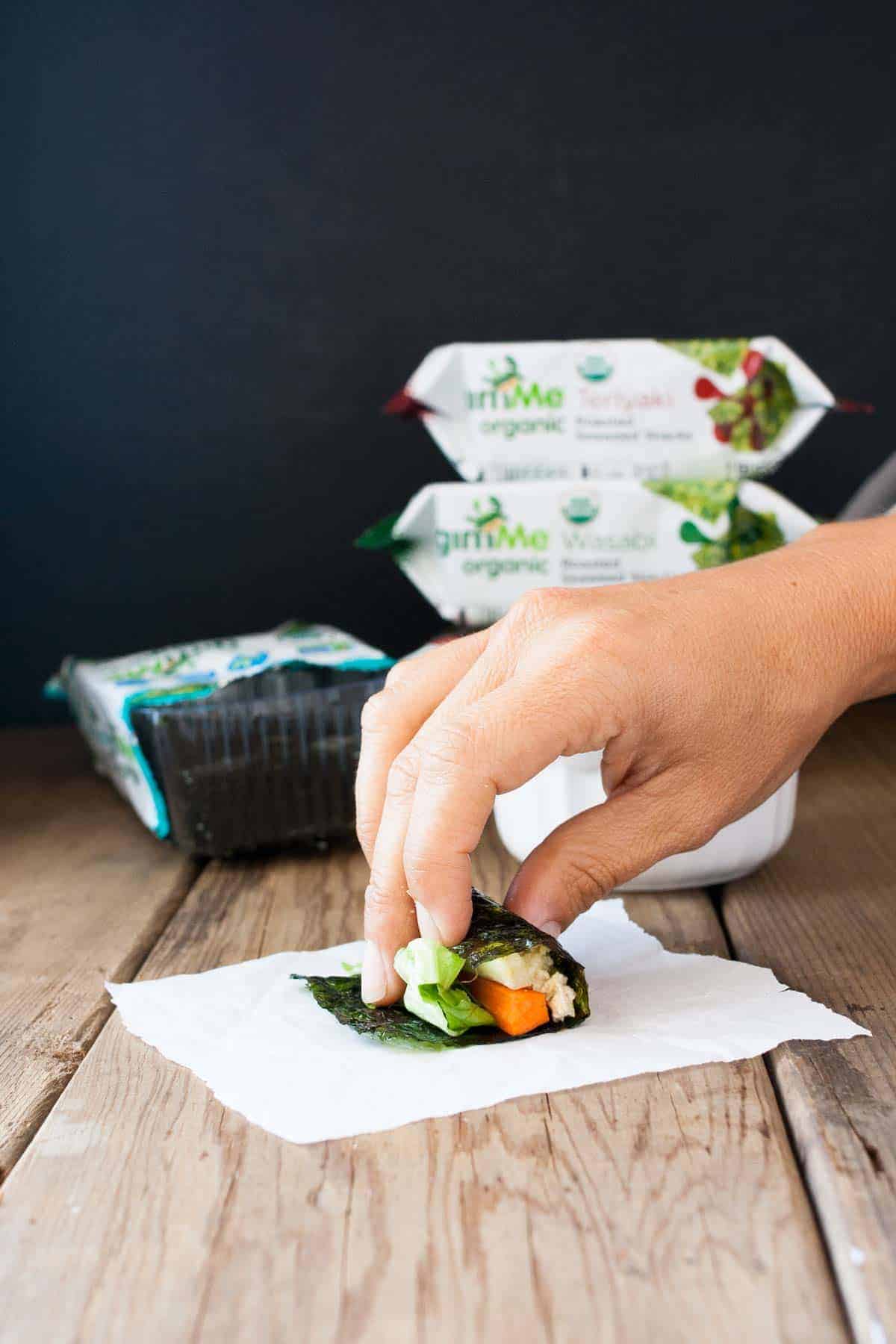 A hand rolling a seaweed snack filled with veggies and rice on top of a paper towel sitting on a wooden surface.