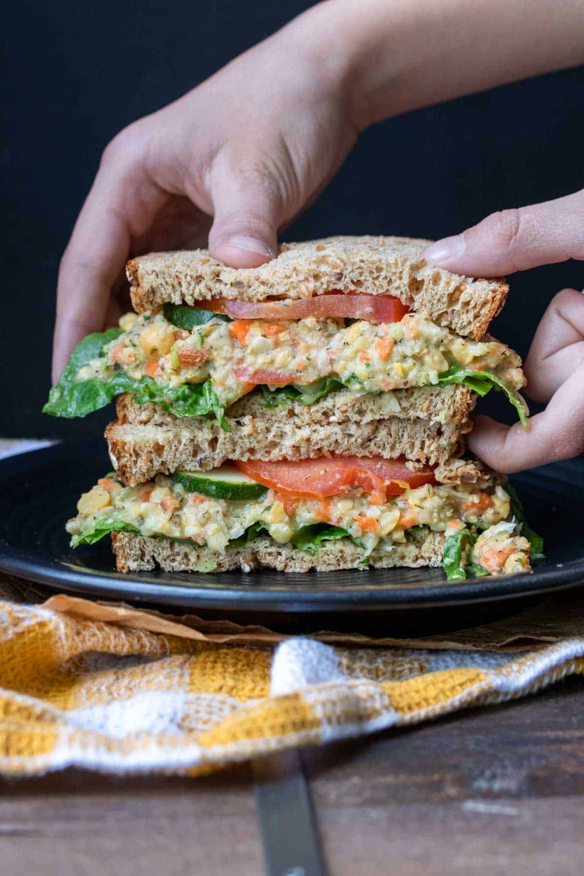 Hands picking up half a mashed chickpea sandwich with veggies.