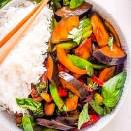 Thai basil eggplant in a white bowl, served with rice and chopsticks on the side.