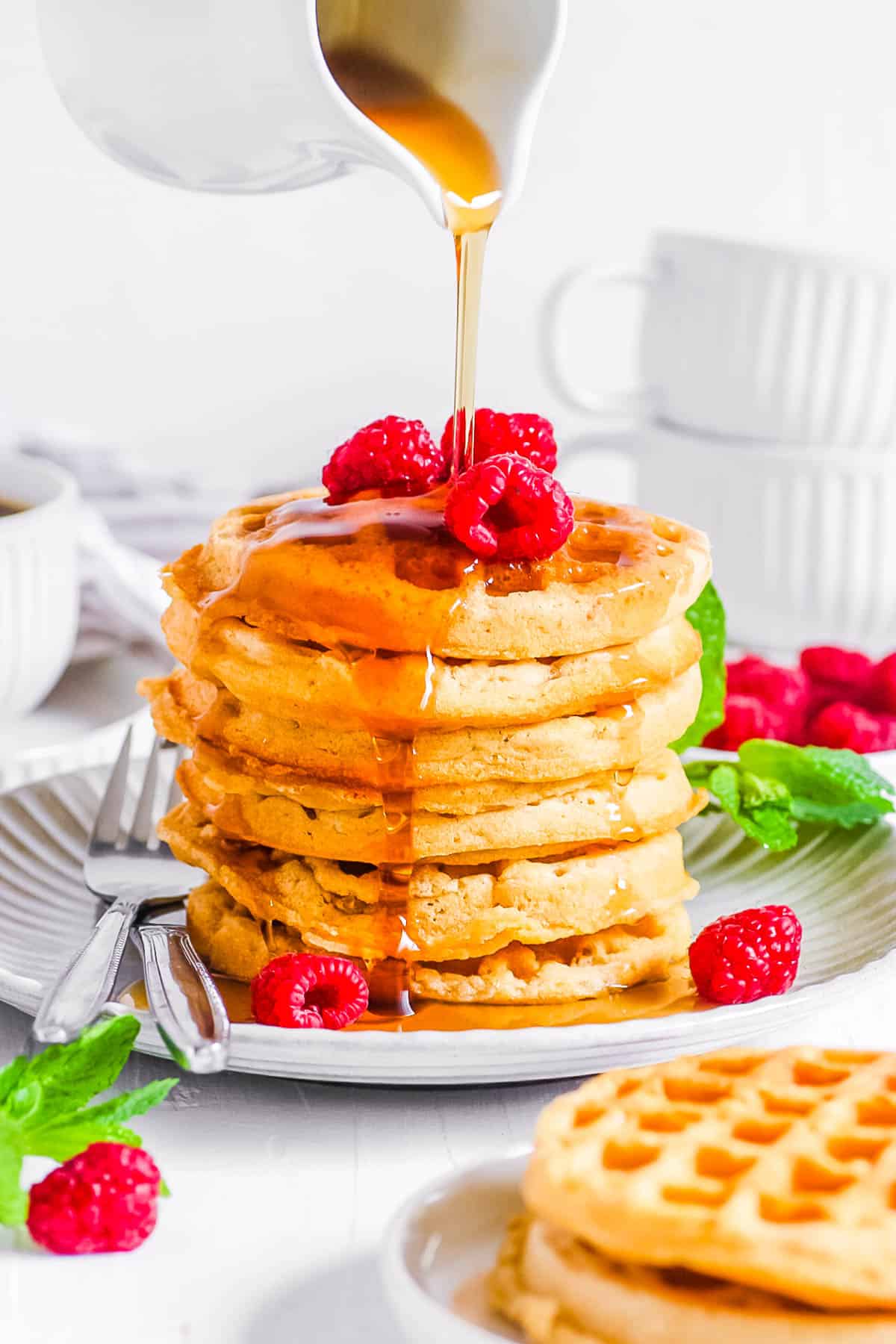 Gluten free vegan waffles stacked on a white plate, topped with raspberries and maple syrup.