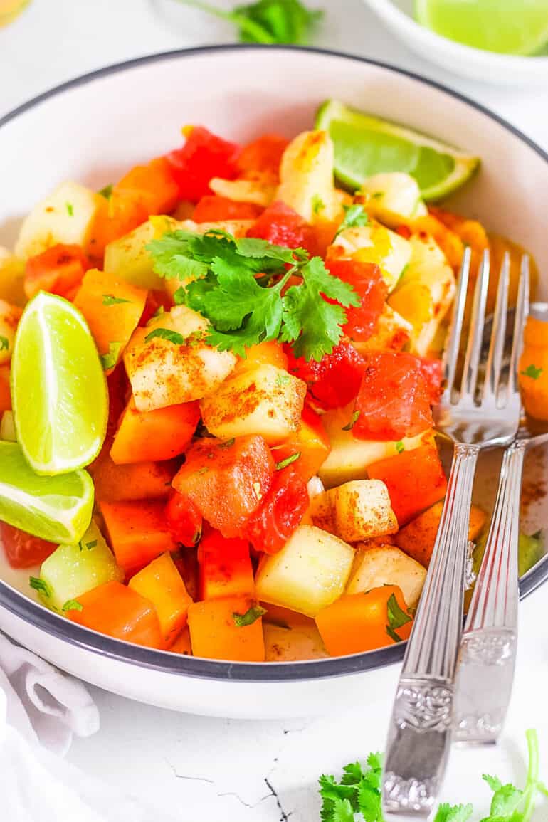 Mexican fruit salad with tajin seasoning, cilantro and fresh lime served in a bowl with a fork.