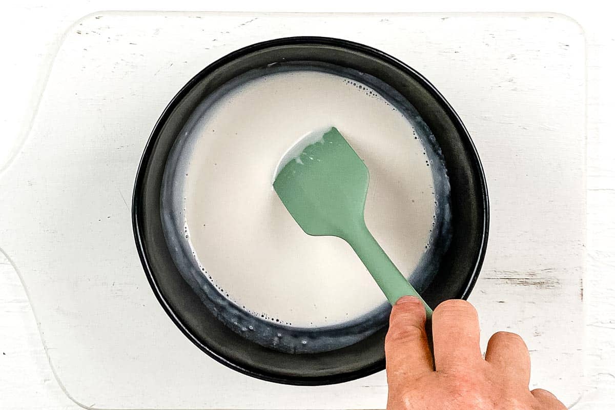 Making the chocolate frosting by warming coconut cream in a mixing bowl on a white background. 