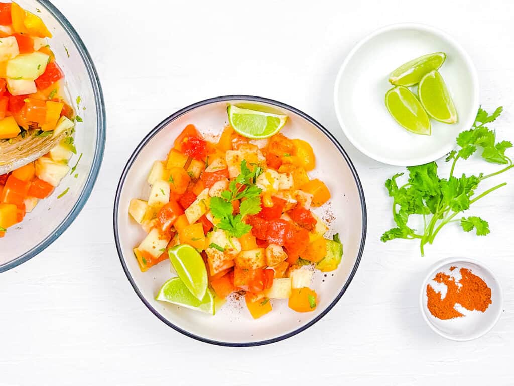 Mexican fruit cup recipe in a white bowl, garnished with cilantro and lime wedges.