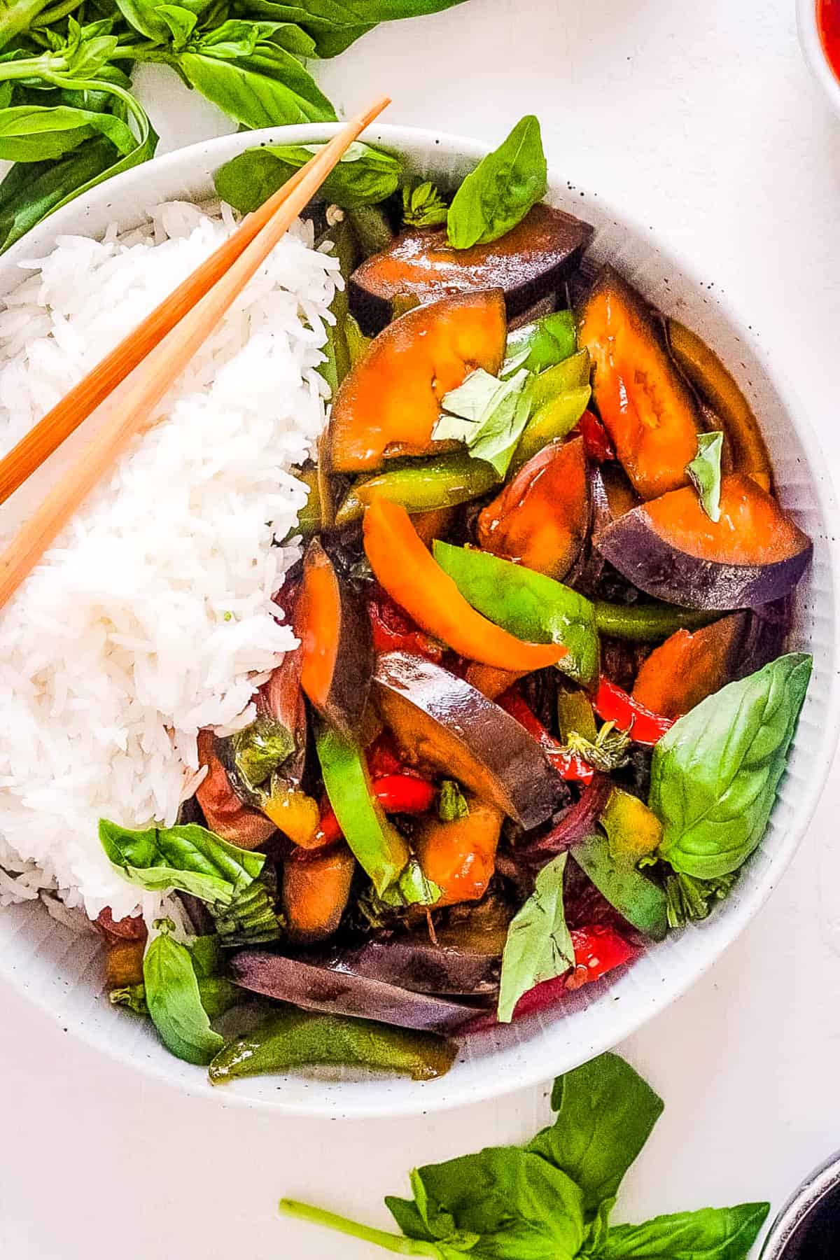 Thai basil eggplant in a white bowl, served with rice and chopsticks on the side.