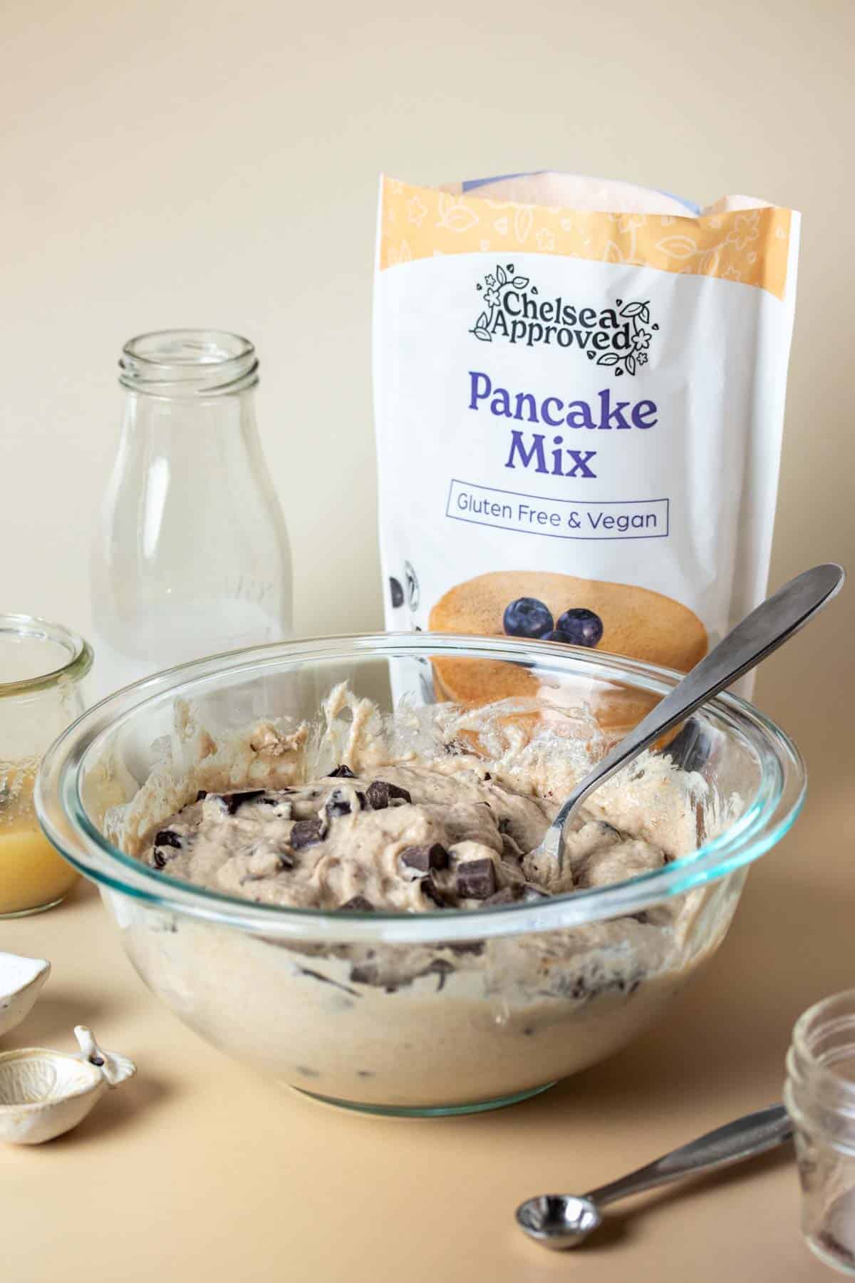 Chocolate chips being mixed into a glass bowl of batter in front of a bag of pancake mix and other ingredients.