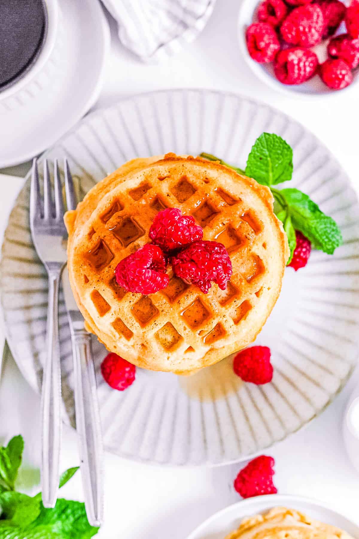 Gluten free vegan waffles stacked on a white plate, topped with raspberries and maple syrup.