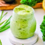 Avocado green goddess dressing in a glass jar surrounded by fresh green herbs.