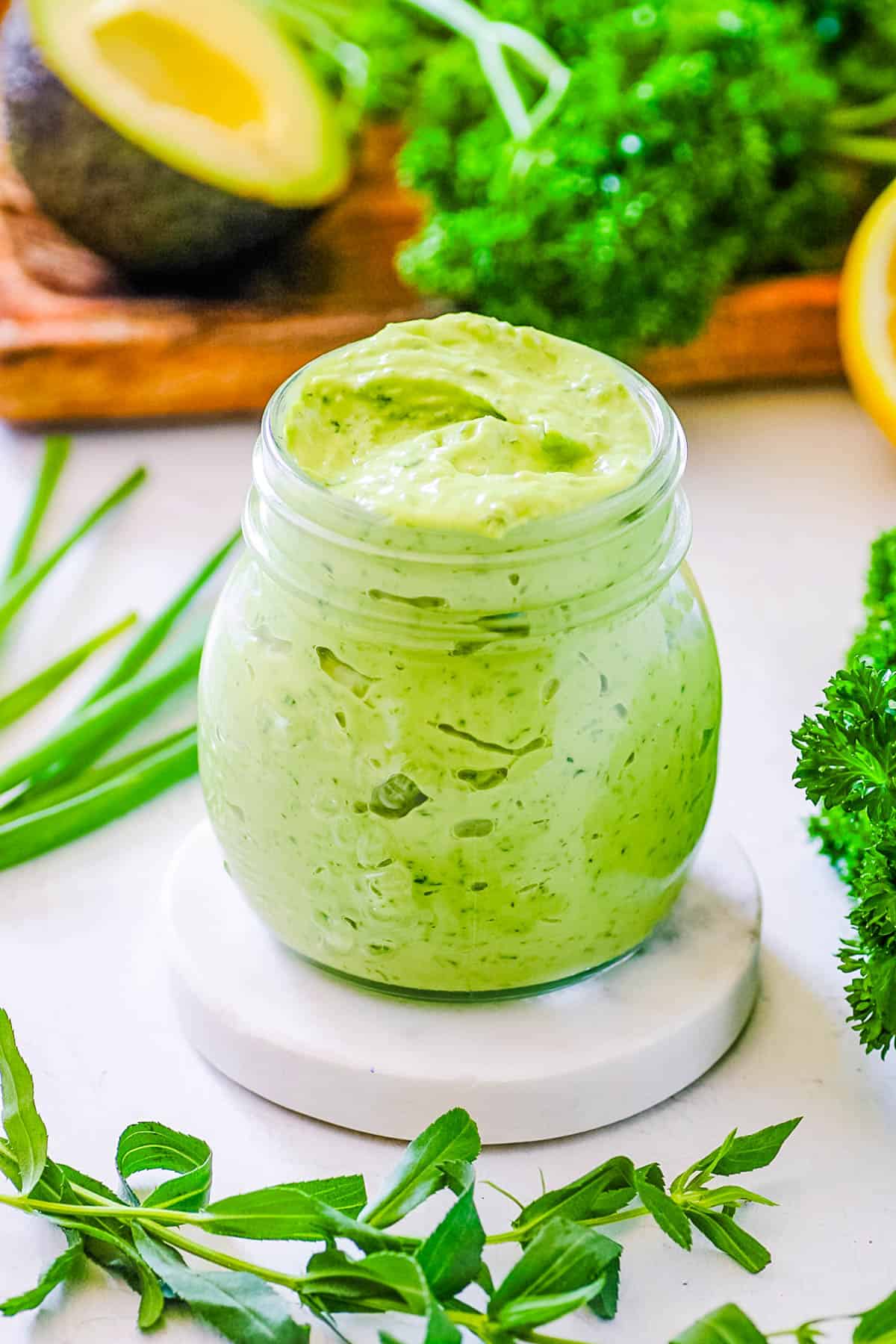 Avocado green goddess dressing in a glass jar surrounded by fresh green herbs.