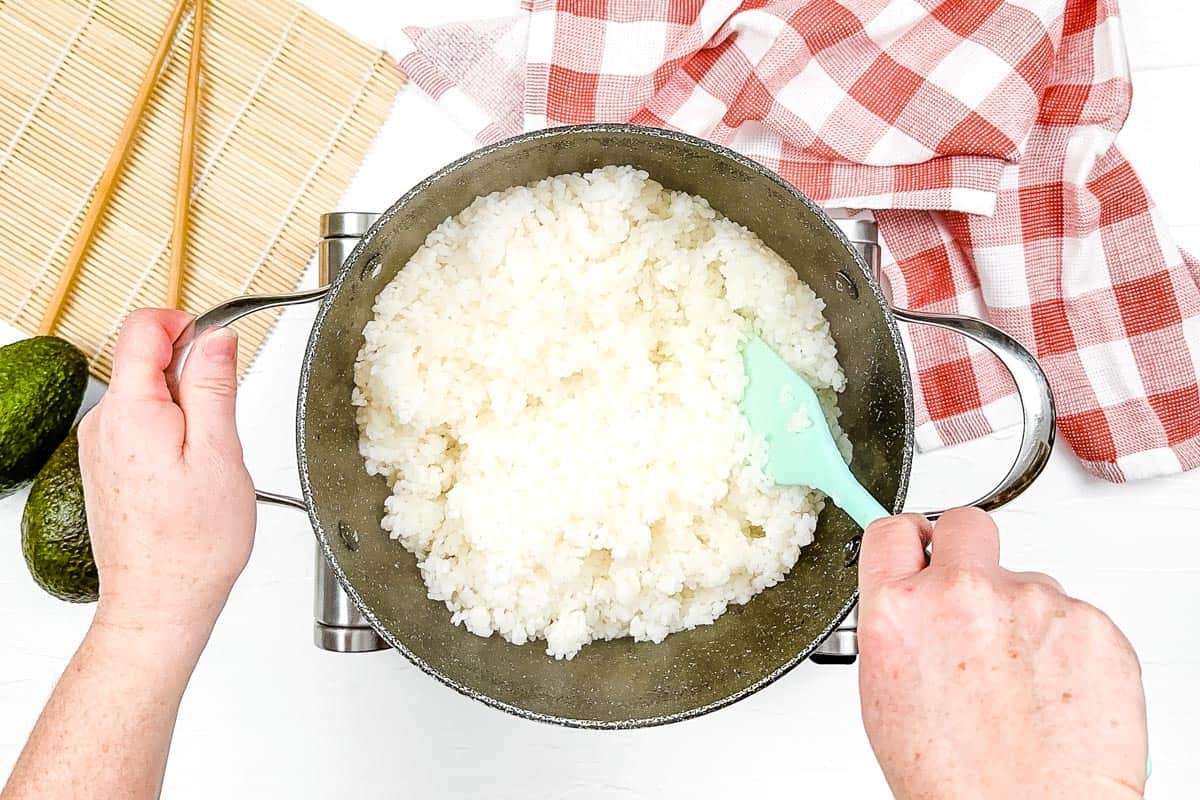 Cooked sushi rice in a large black pot being mixed with a green spatula.