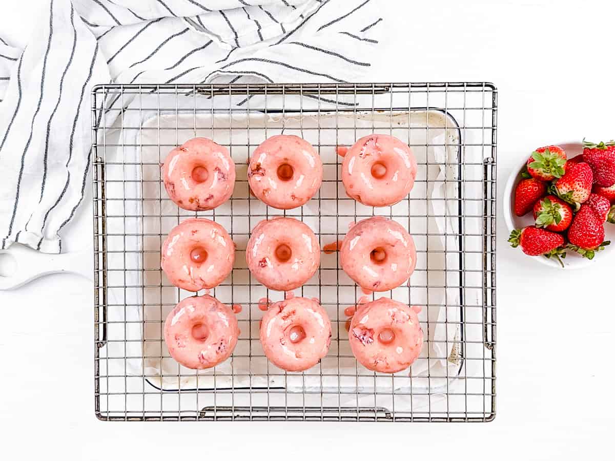 Baked strawberry donuts glazed and sitting on a wire rack so the glaze can drip off, beside a bowl of fresh strawberries.