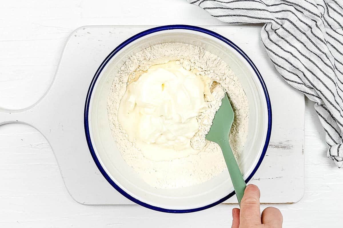 Wet ingredients are mixed into the dry ingredients with a spatula, on a white background. 