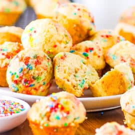 Birthday cake muffins, stacked on a white plate with sprinkles on the side.