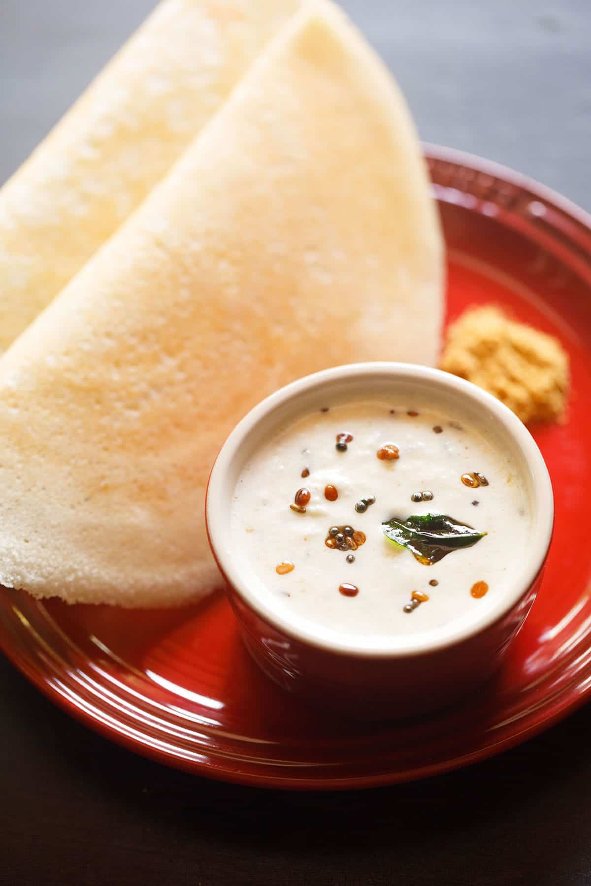 a small white bowl of coconut chutney on a red plate with two dosa