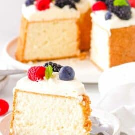 A slice of angel food cake on a white plate with whipped cream and fresh berries and a spoon.
