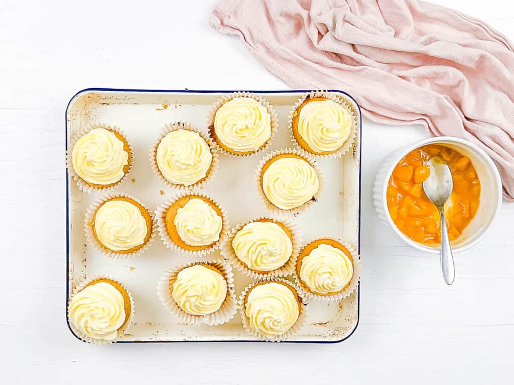 Peach filled cupcakes topped with a buttercream frosting on a baking sheet.