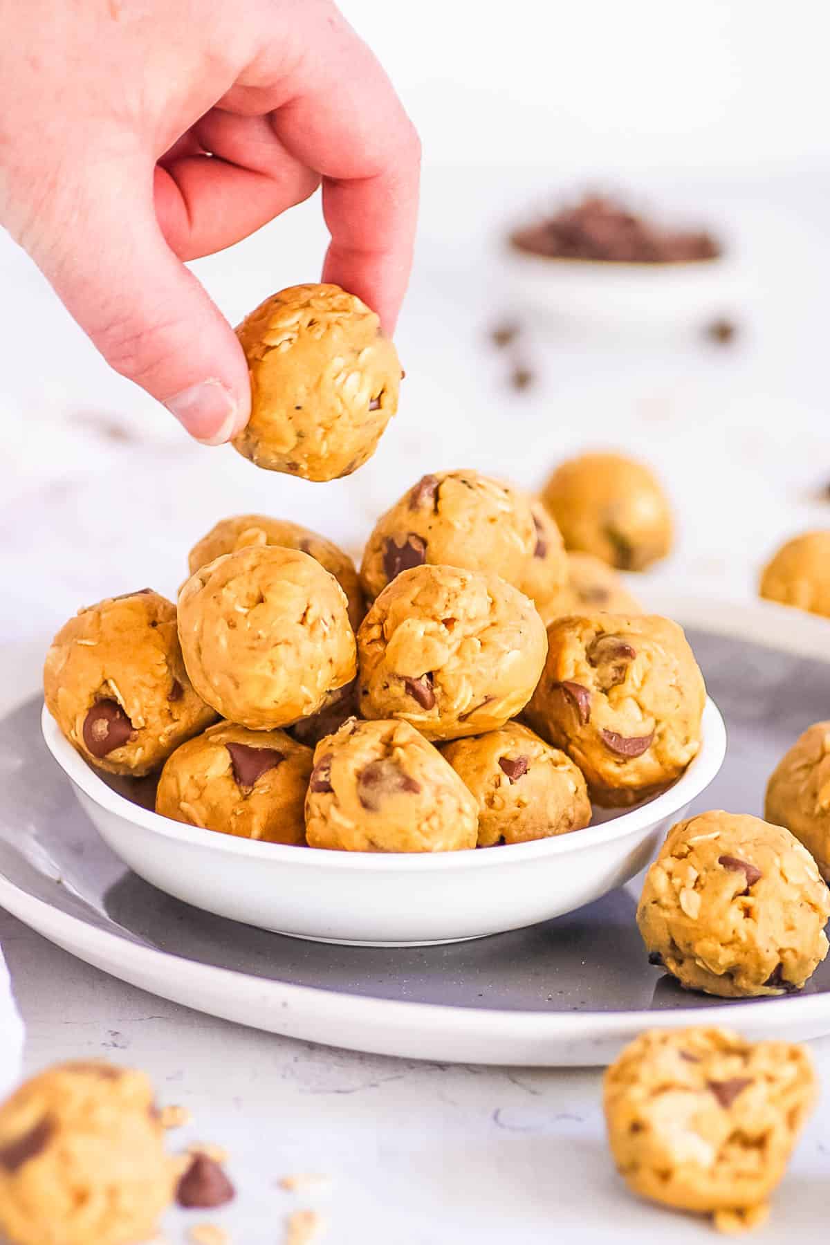 Stack of vegan protein balls made with peanut butter, oats, protein powder, and chocolate chips, in a white bowl with a hand picking up one ball.