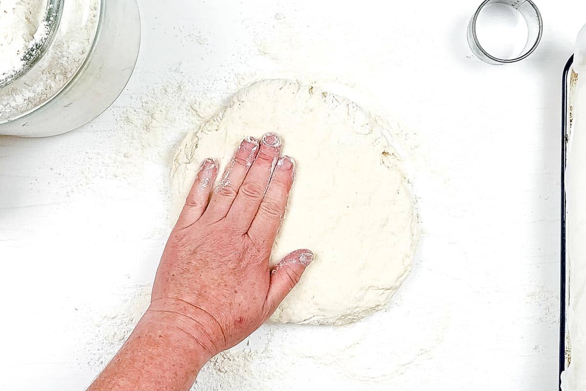 A hand is shaping the biscuit dough into a rough circle before cutting it. 