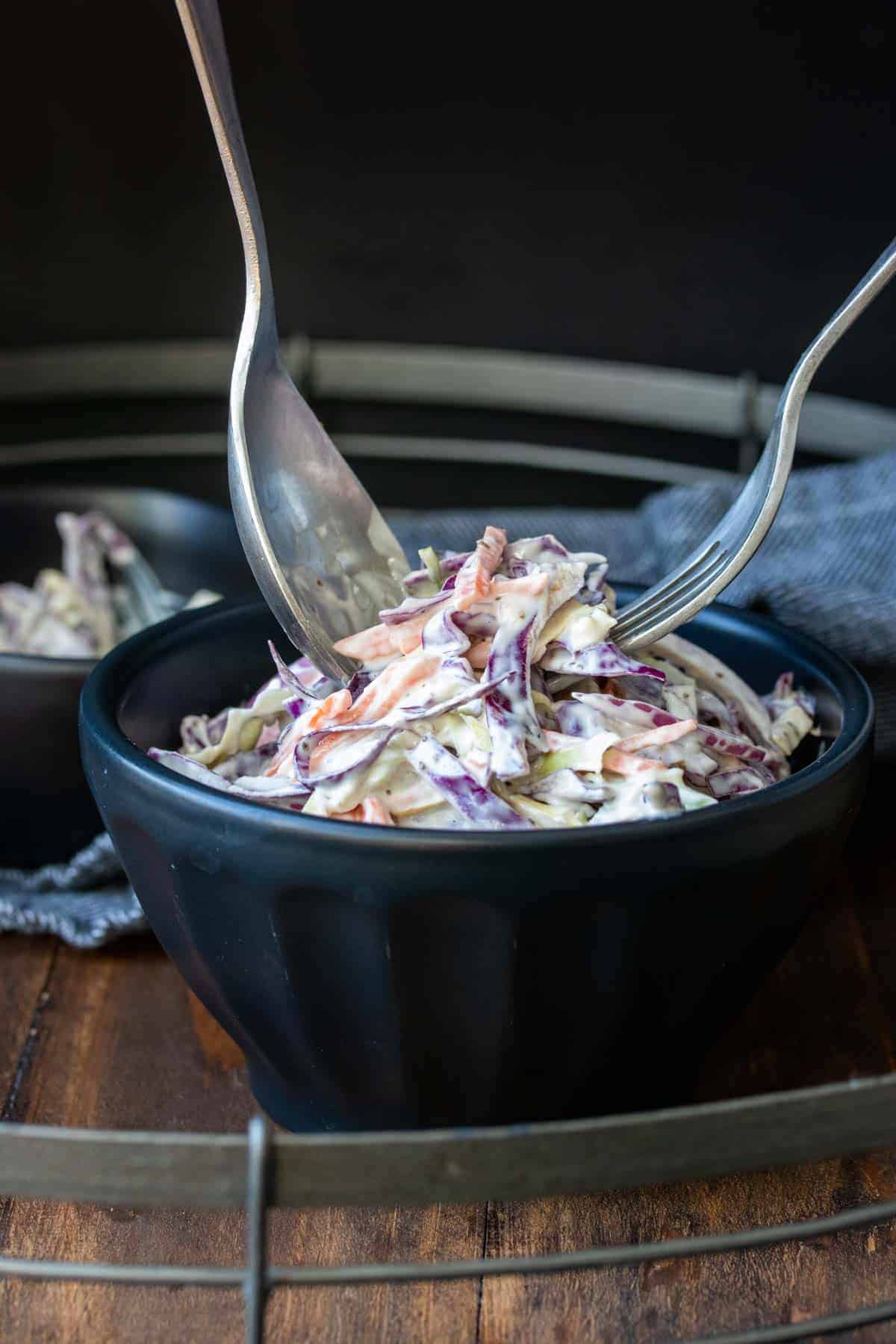 Two forks getting creamy coleslaw out of a black bowl.