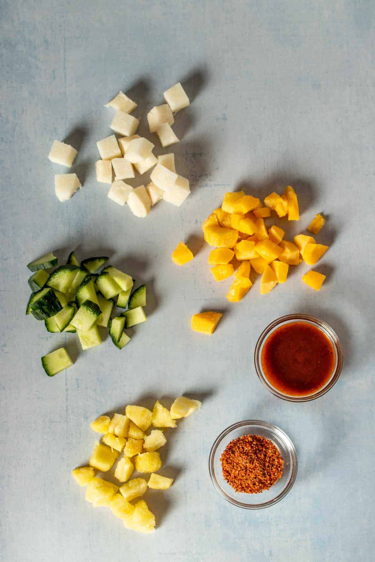 Top view of piles of chopped cucumber, jicama, mango, pineapple and small glass bowls of a red sauce and chile lime seasoning.