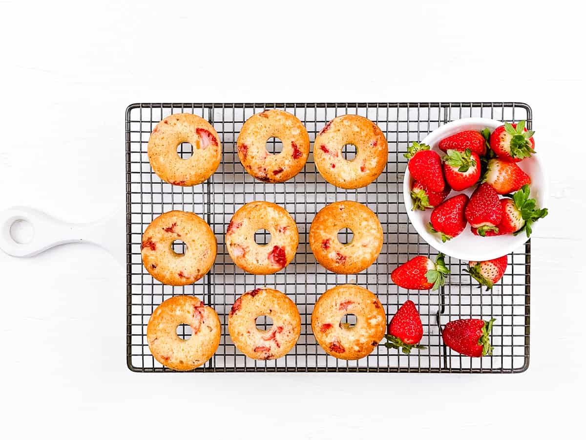 Baked strawberry donuts being cooled on a cooling rack with bowl of fresh strawberries beside them.