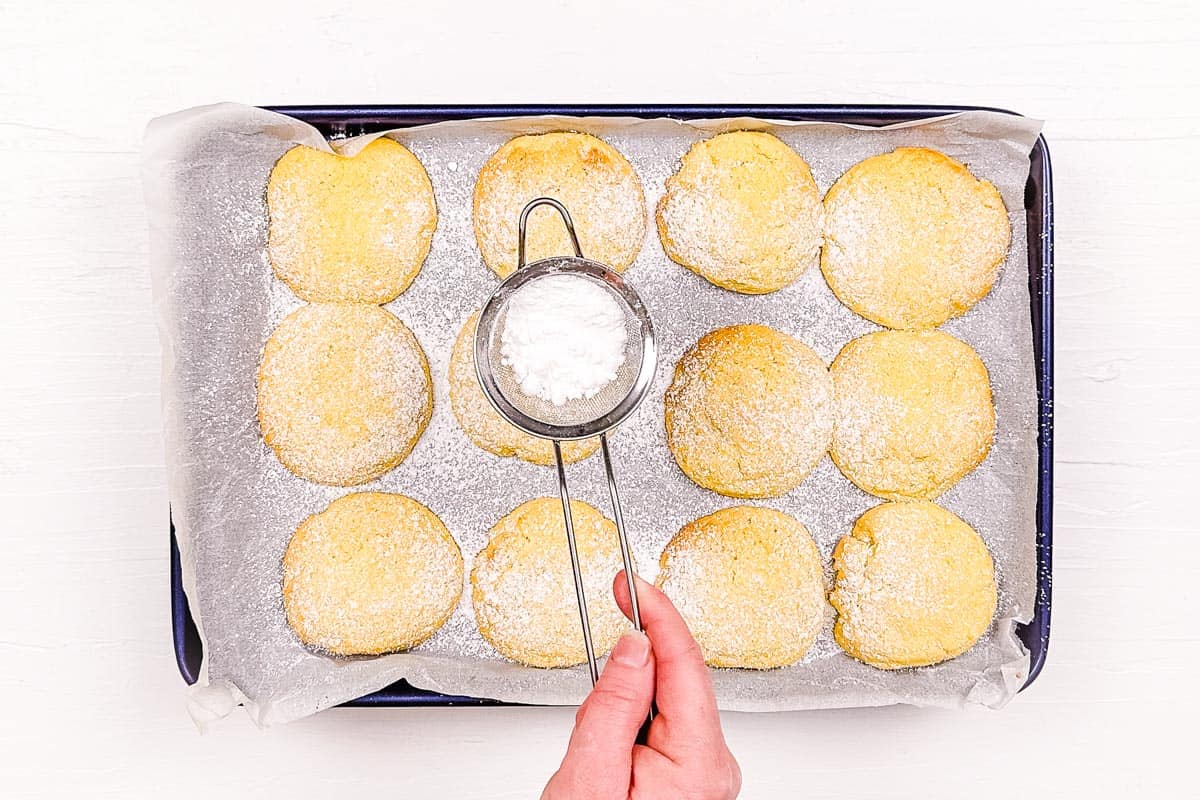 Sprinkling powdered sugar over the baked lemon biscuits using a small sieve.