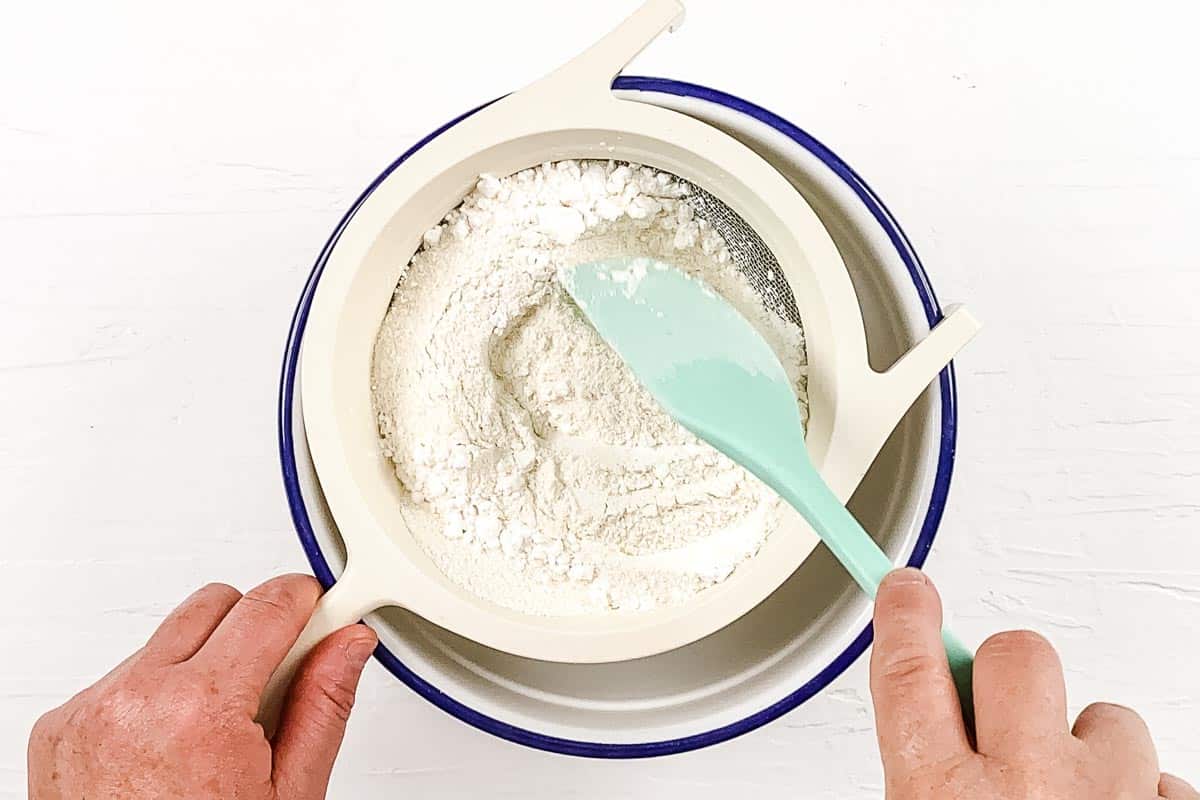 Sifting the dry ingredients into a large mixing bowl using a metal sieve and a green spatula.
