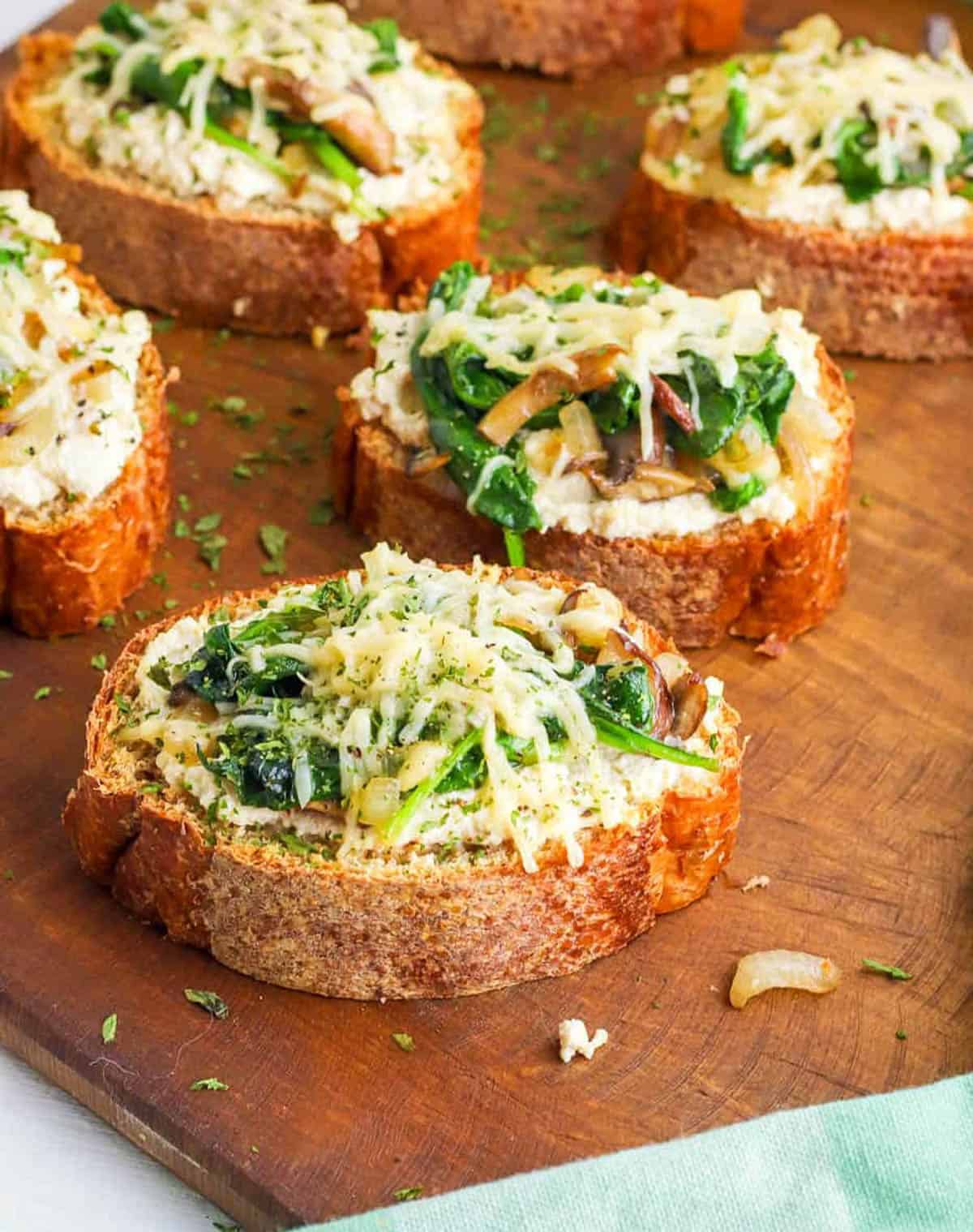 Mushroom toast topped with spinach and parmesan cheese on a wooden cutting board.