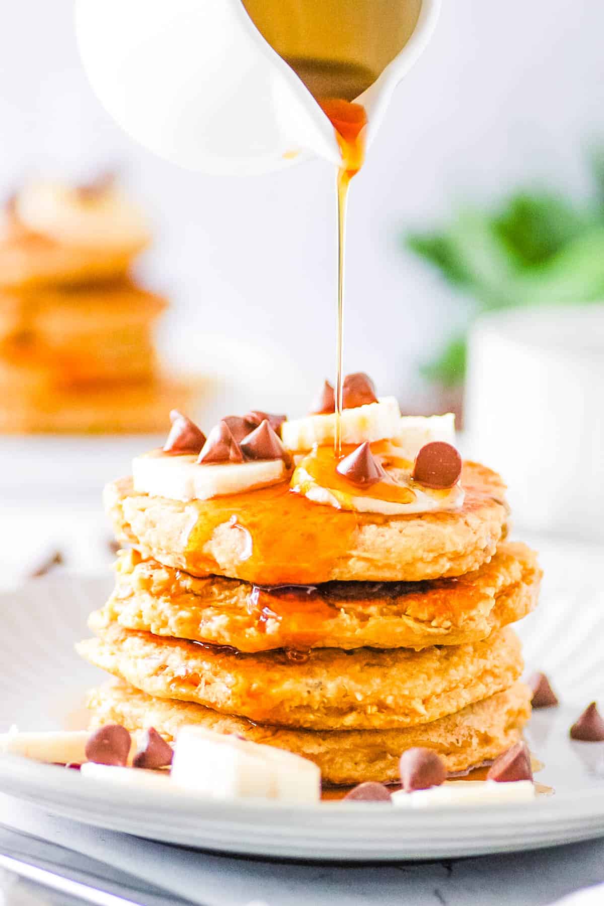 Stack of oat flour pancakes topped with chocolate chips, banana slices and maple syrup on a white plate.