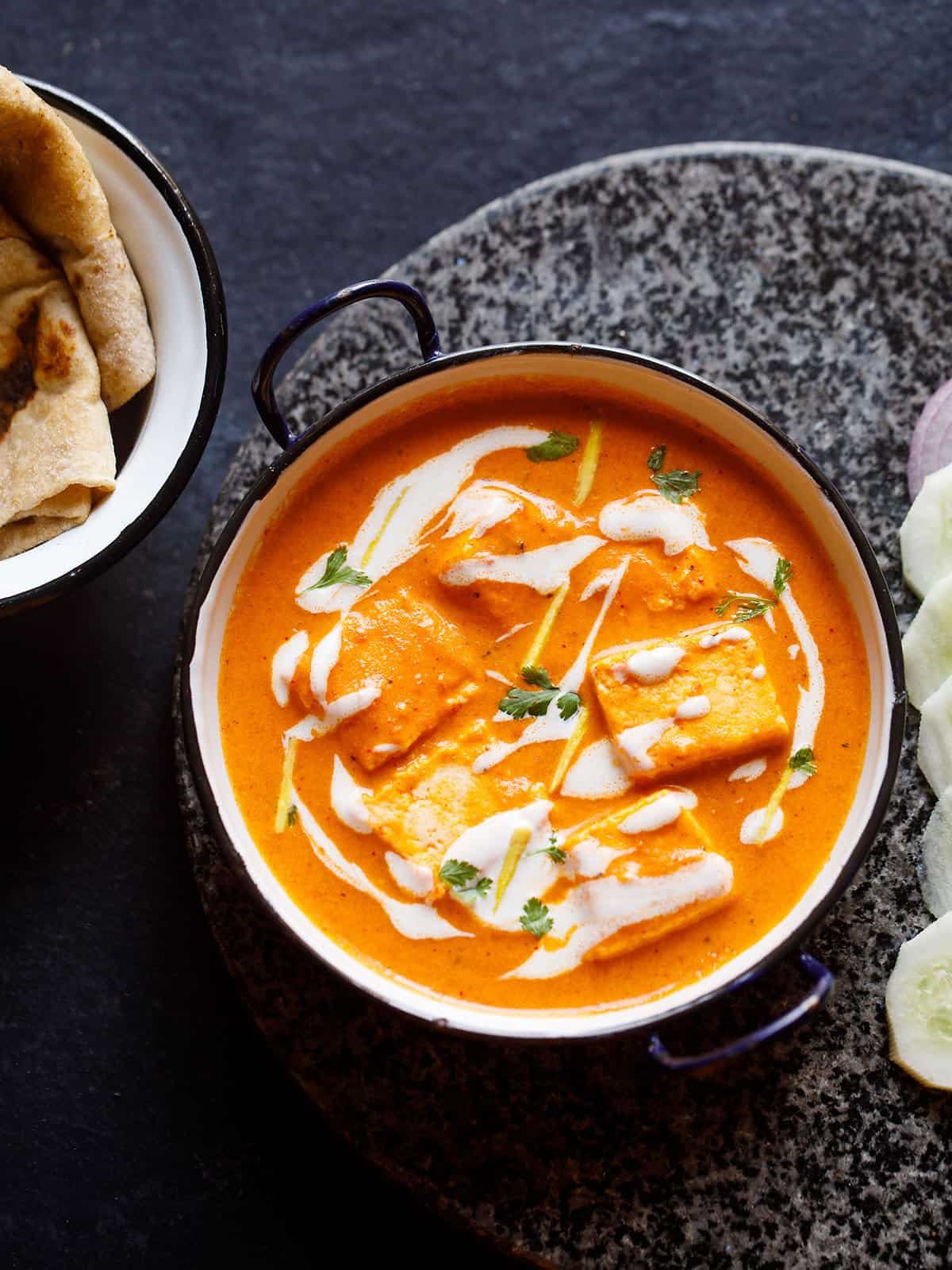 paneer butter masala served in a blue rimmed white pan, garnished with cream and cilantro with a side of sliced cucumber and roti