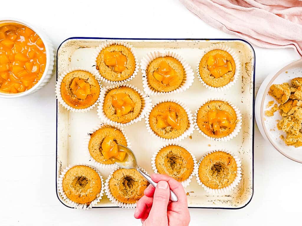 Peach cobbler cupcakes on a baking sheet, with a spoon coring out the centers of the cupcakes.