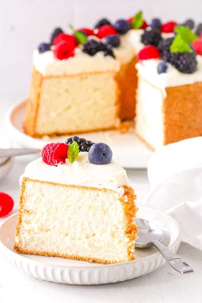 A slice of angel food cake on a white plate with whipped cream and fresh berries and a spoon.