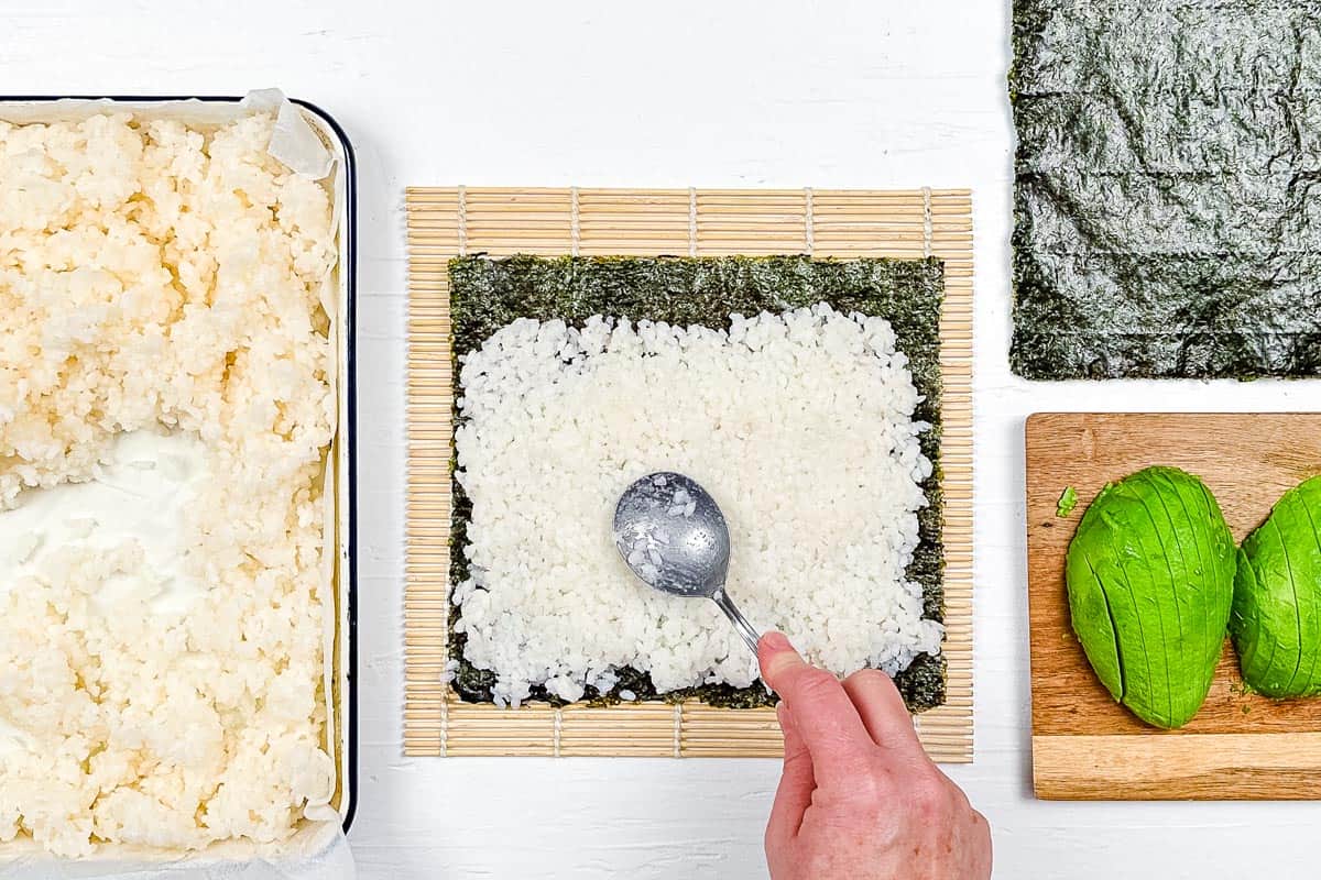 Patting down the sushi rice in an even layer over the seaweed nori before adding the avocado slices.