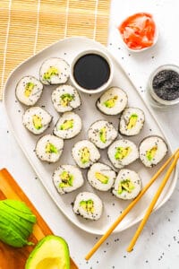 A large white platter with sliced avocado sushi rolls laid out with soy sauce and pink ginger.