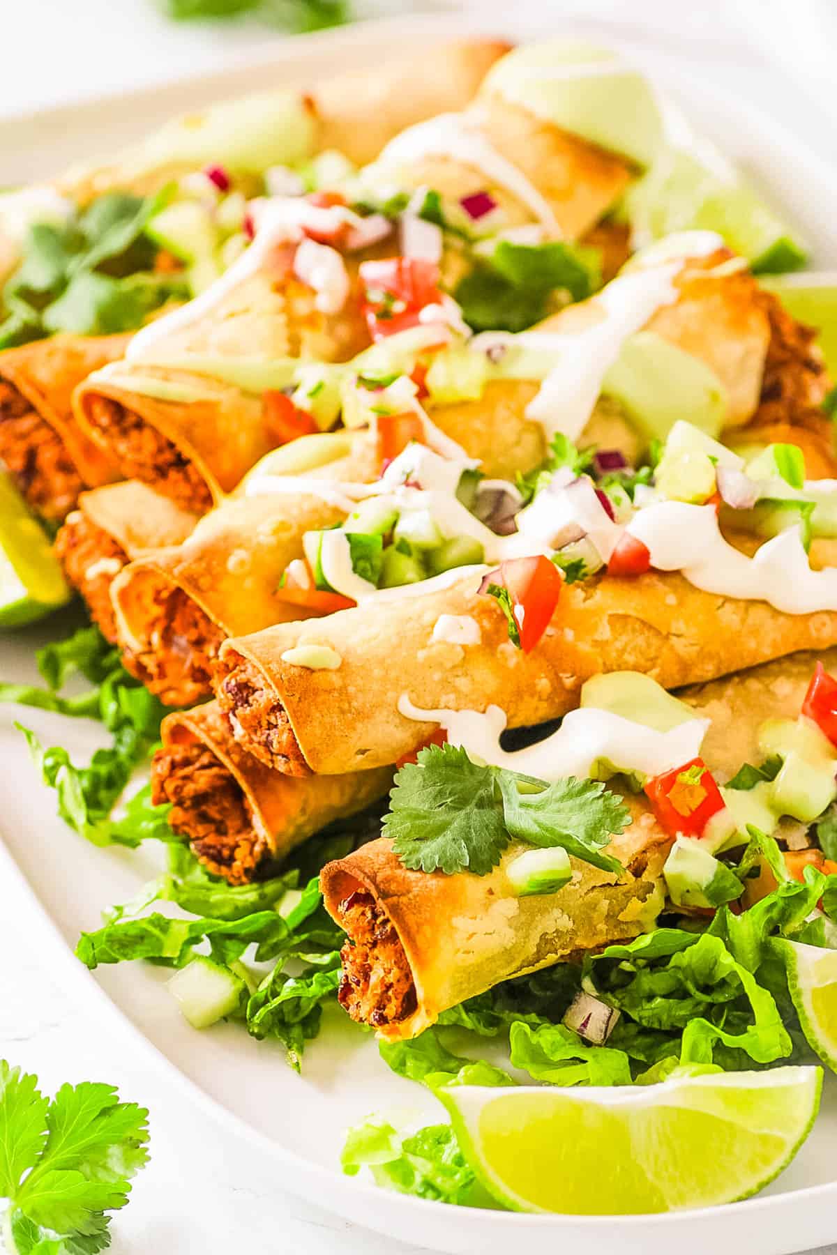 Baked vegan taquitos topped with vegan sour cream, salsa and served on a white platter with lettuce and limes.