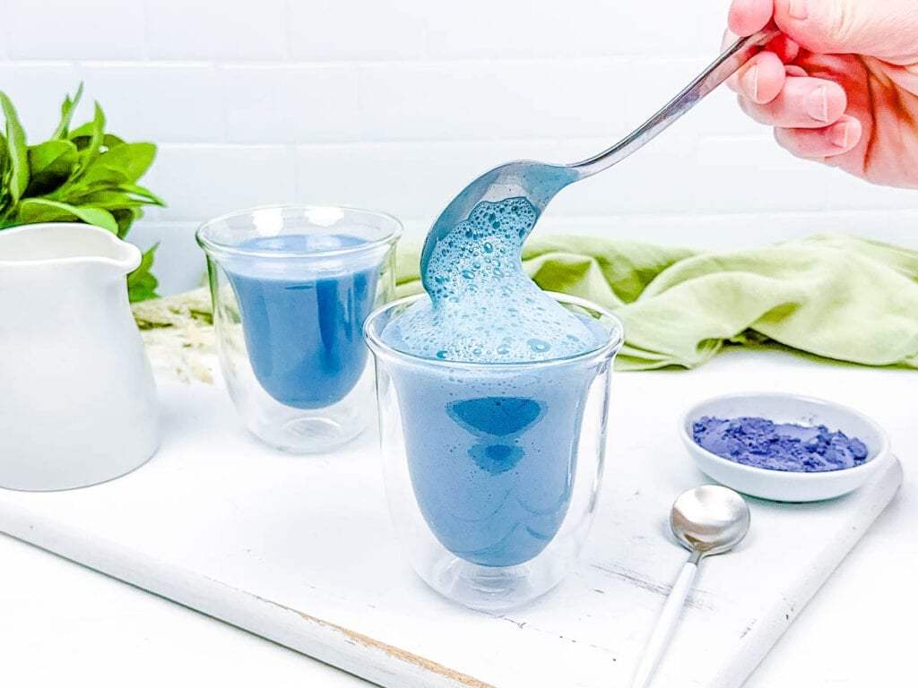 Blue butterfly latte served in a glass, with almond milk foam spooned on top.