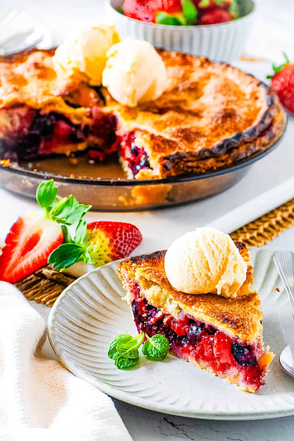 Slice of easy strawberry blueberry pie topped with ice cream with a whole pie in the background.