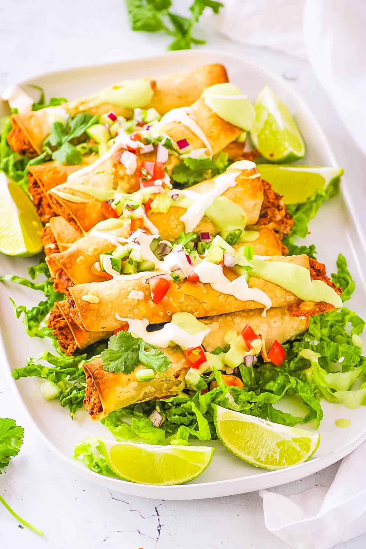 Healthy vegan taquitos topped with vegan sour cream, salsa and served on a white platter with lettuce and limes.
