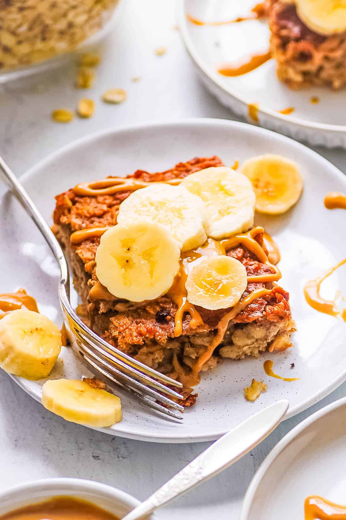 One slice of banana peanut butter baked oatmeal, garnished with banana slices, served on a white plate with a fork.