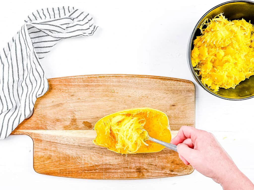 Cooked spaghetti squash with squash "strings" being pulled out with a fork on a cutting board.
